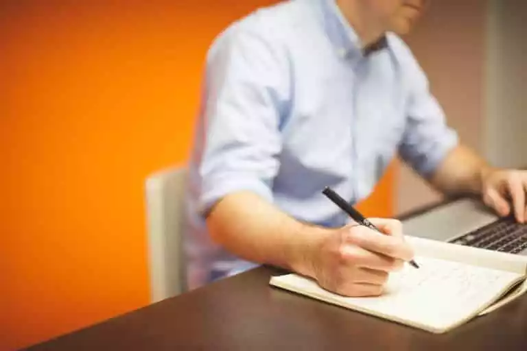 Un homme écrit une publicité pour une caution sur un carnet devant un mur orange.