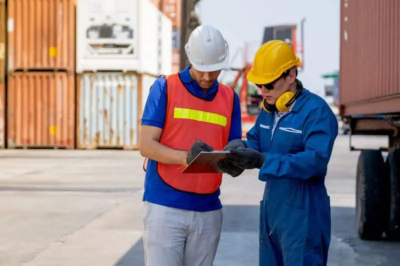 Cargo container worker and engineer discuss together with data in document on clipboard