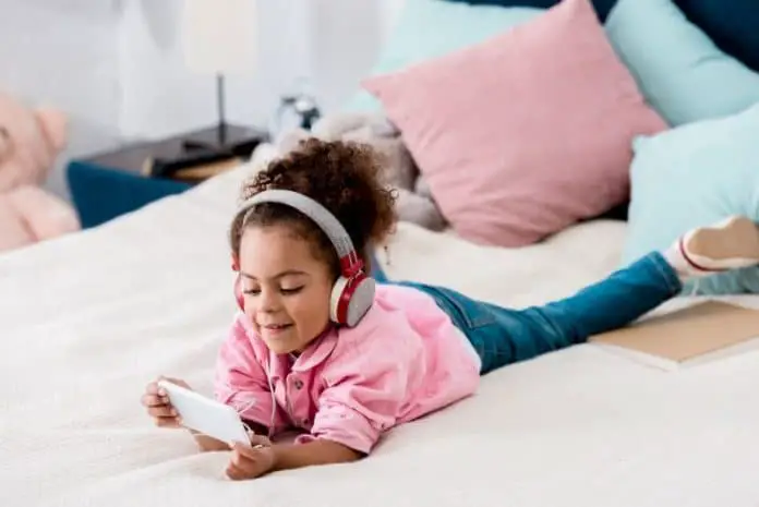 cute african american child lying on the bed and listening music on her smartphone in headphones