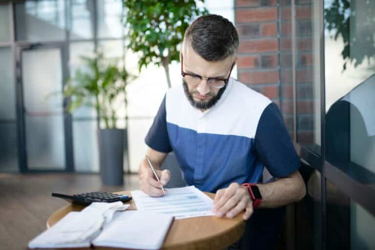 Un homme portant des lunettes et une barbe est assis à une table concentré sur le remplissage d un formulaire avec un stylo une calculatrice et des documents sont étalés sur la table