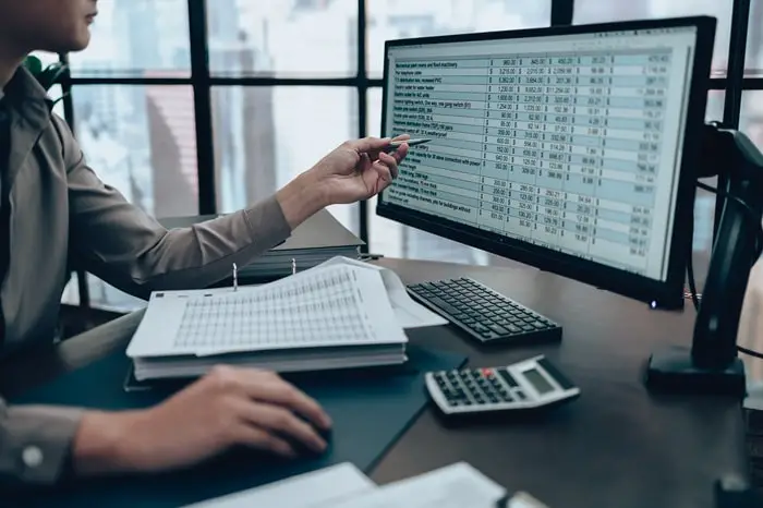 Personne travaillant à un bureau avec des feuilles de calcul financières sur un écran d'ordinateur, tenant un stylo, avec des documents ouverts et une calculatrice à proximité.