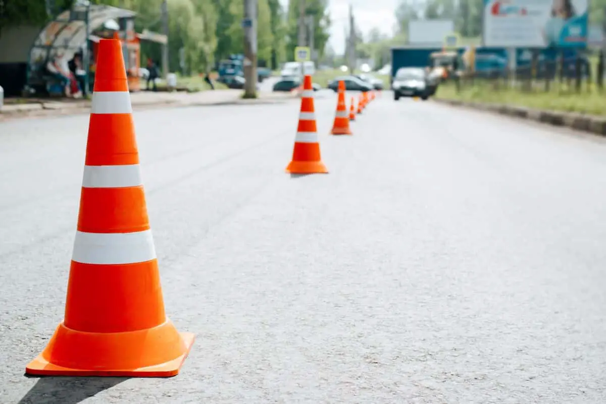 Des cônes de signalisation orange alignés sur une route grise avec des véhicules flous et de la verdure en arrière plan