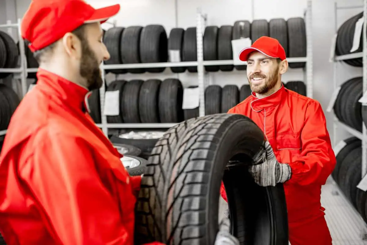 Deux ouvriers en uniformes rouges manipulent un pneu de voiture dans un entrepôt dont les étagères sont remplies de pneus
