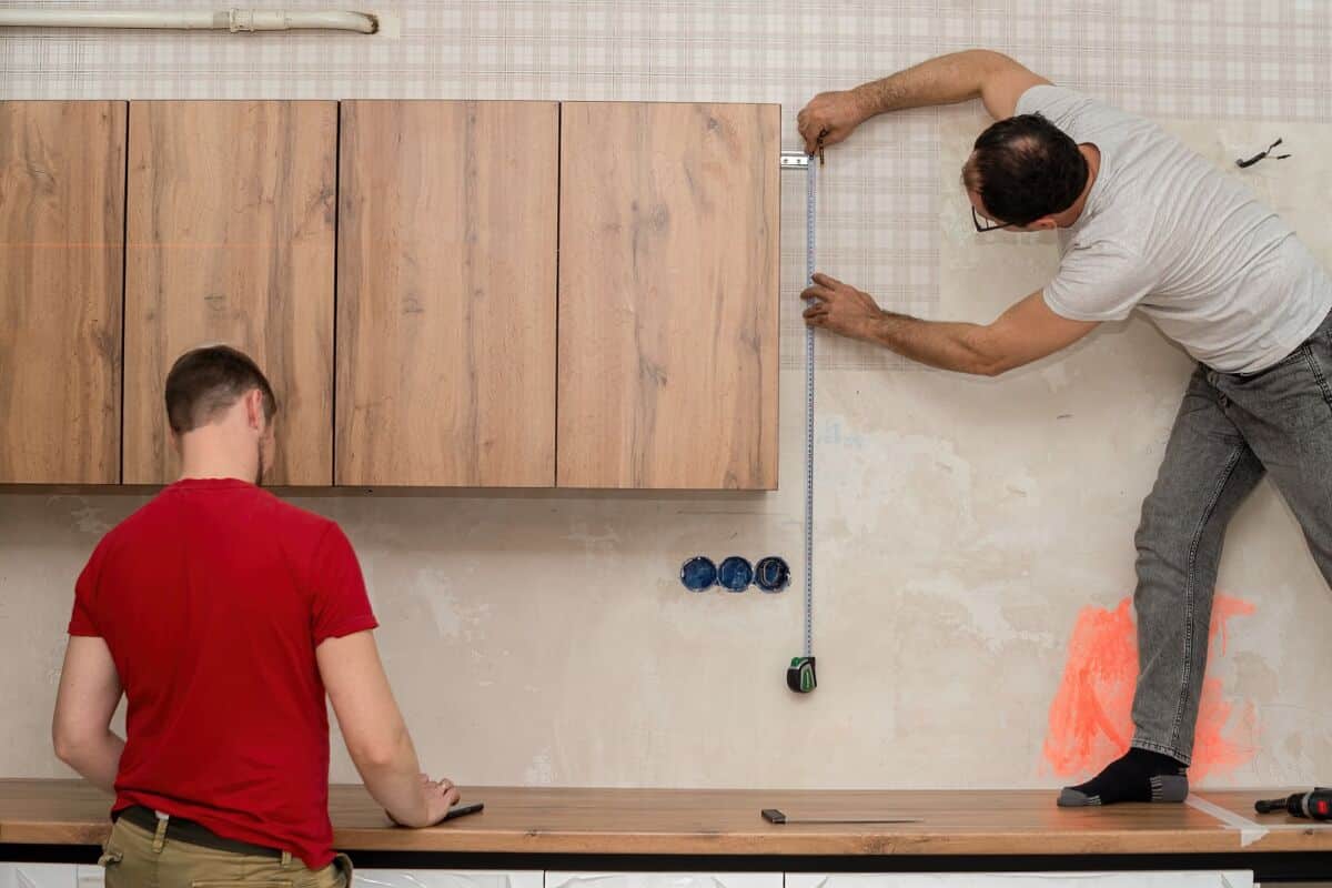 Deux hommes travaillent dans une cuisine : l un mesure un mur avec un mètre ruban tout en se tenant sur le comptoir l autre prépare du matériel sur le plan de travail
