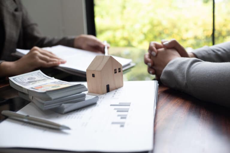 Deux personnes à une table discutent de documents. On peut voir une petite maison en bois, des piles de billets et un graphique.