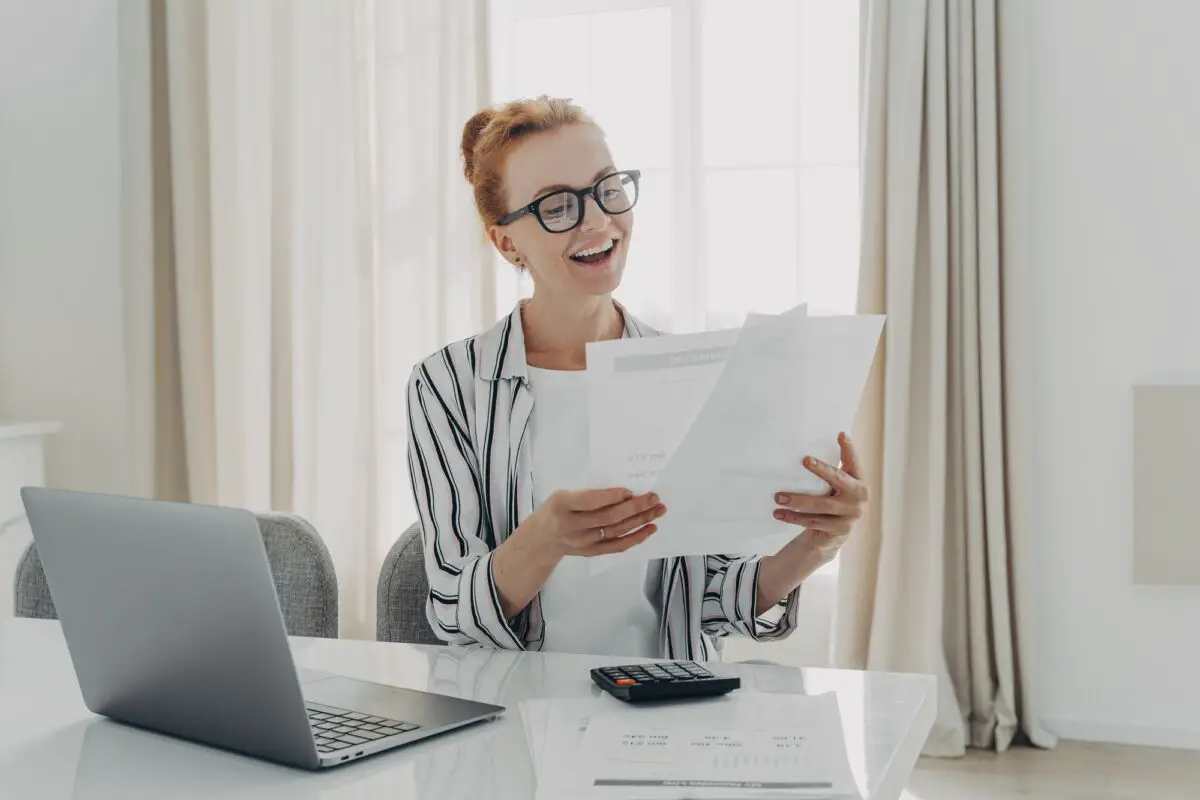 Une femme assise à un bureau avec un ordinateur portable et une calculatrice souriant tout en tenant et en regardant plusieurs documents