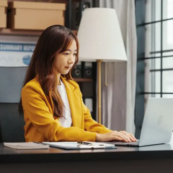 Personne assise à un bureau travaillant sur un ordinateur portable dans un bureau bien éclairé avec des papiers et une lampe à proximité