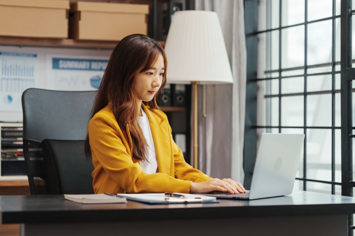 Personne assise à un bureau travaillant sur un ordinateur portable dans un bureau bien éclairé avec des papiers et une lampe à proximité