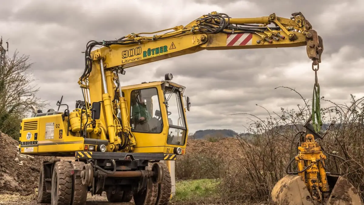 Excavatrice jaune avec une griffe sur un chantier de construction extérieur, entourée de terre et de buissons sous un ciel nuageux.