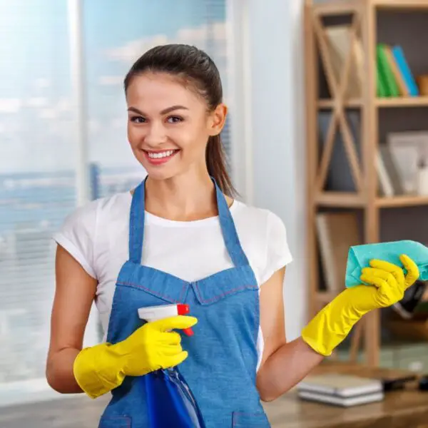 Personne portant un tablier bleu et des gants jaunes tenant un vaporisateur et un chiffon, debout dans une pièce propre avec une étagère en arrière-plan.