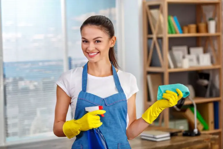 Personne portant un tablier bleu et des gants jaunes tenant un vaporisateur et un chiffon, debout dans une pièce propre avec une étagère en arrière-plan.