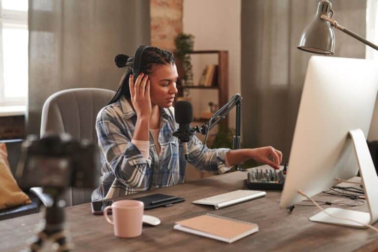 Une personne portant un casque ajuste le mixeur audio devant un ordinateur et un microphone assise à un bureau avec une caméra une tasse et un bloc notes