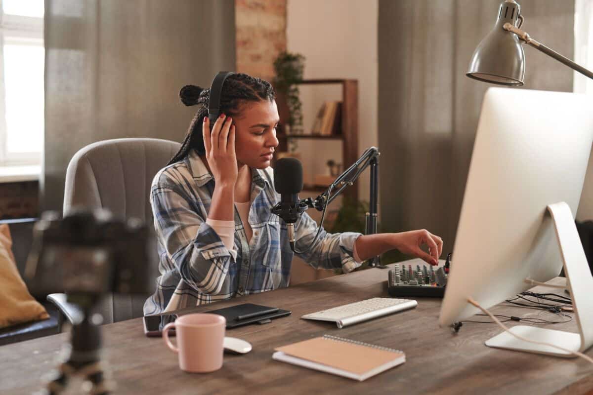 Une personne portant un casque ajuste le mixeur audio devant un ordinateur et un microphone assise à un bureau avec une caméra une tasse et un bloc notes