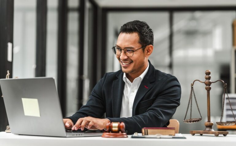 Un homme en costume sourit en tapant sur un ordinateur portable dans un bureau. Un marteau et une balance de la justice sont posés sur le bureau.