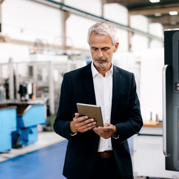 Un homme âgé en costume examine une tablette dans une usine, avec des machines et des équipements industriels en arrière-plan.