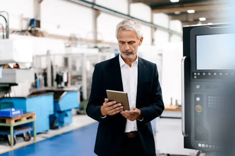Un homme âgé en costume examine une tablette dans une usine, avec des machines et des équipements industriels en arrière-plan.