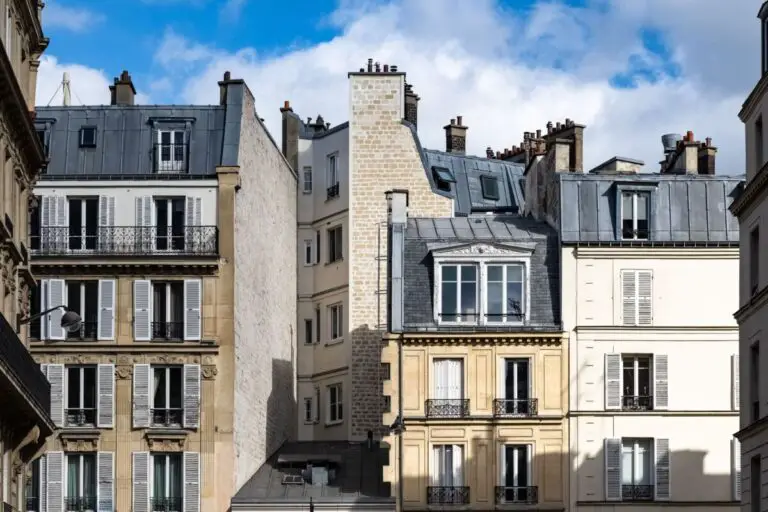 Des immeubles d'appartements à l'architecture parisienne classique, avec balcons et toits mansardés, sous un ciel bleu partiellement nuageux.