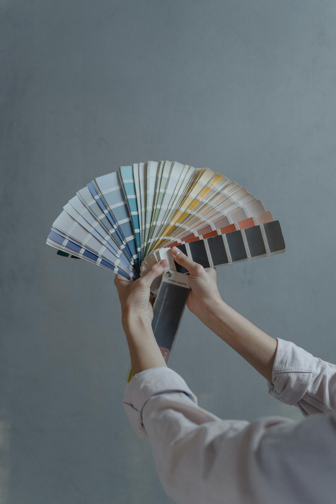 Mains tenant un livret d'échantillons de couleurs de peinture en éventail sur un fond gris.