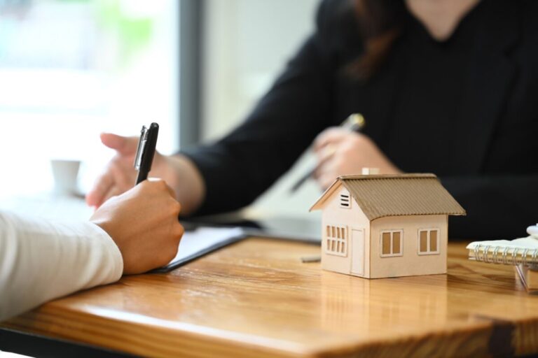 Deux personnes sont assises à une table l une d elles signe un document une petite maquette de maison en bois est posée sur la table