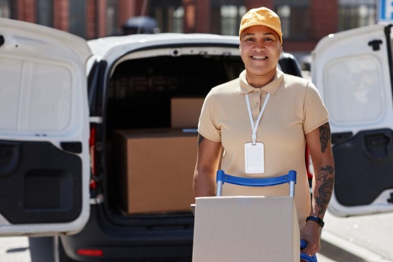 Une personne portant une casquette jaune et une chemise beige se tient devant une camionnette, tenant une boîte sur un diable.