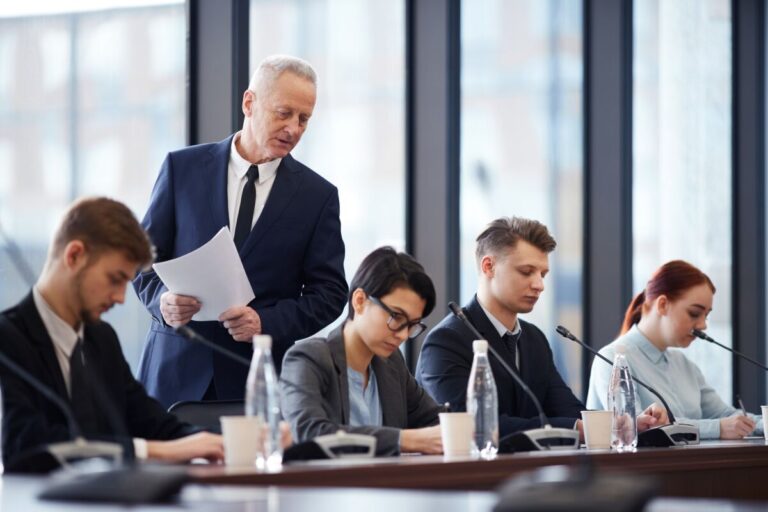 Un homme âgé en costume tenant des documents se tient debout tandis que quatre personnes assises à une table travaillent sur des documents des bouteilles d eau et des microphones sont posés sur la table dans une salle de conférence