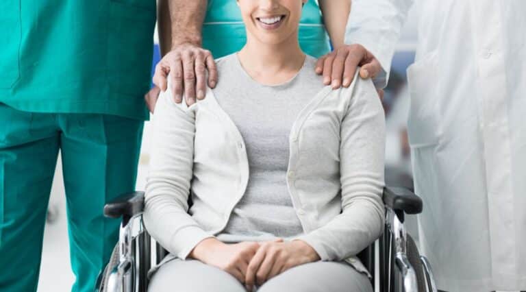 Une personne souriante assise dans un fauteuil roulant est flanquée de deux professionnels de la santé