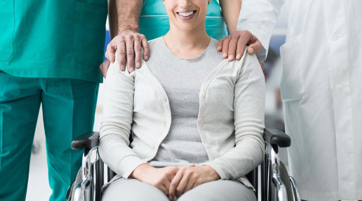 Une personne souriante assise dans un fauteuil roulant est flanquée de deux professionnels de la santé