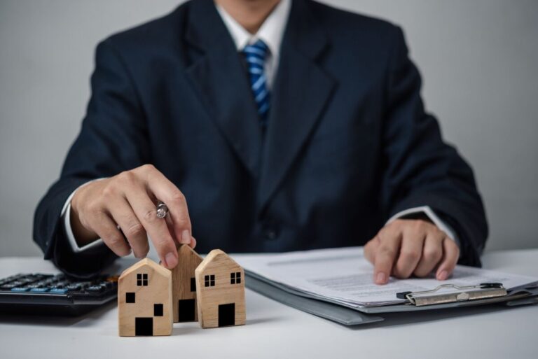 Une personne en costume est assise à un bureau avec des modèles de maisons en bois une calculatrice et des documents sur un presse papiers