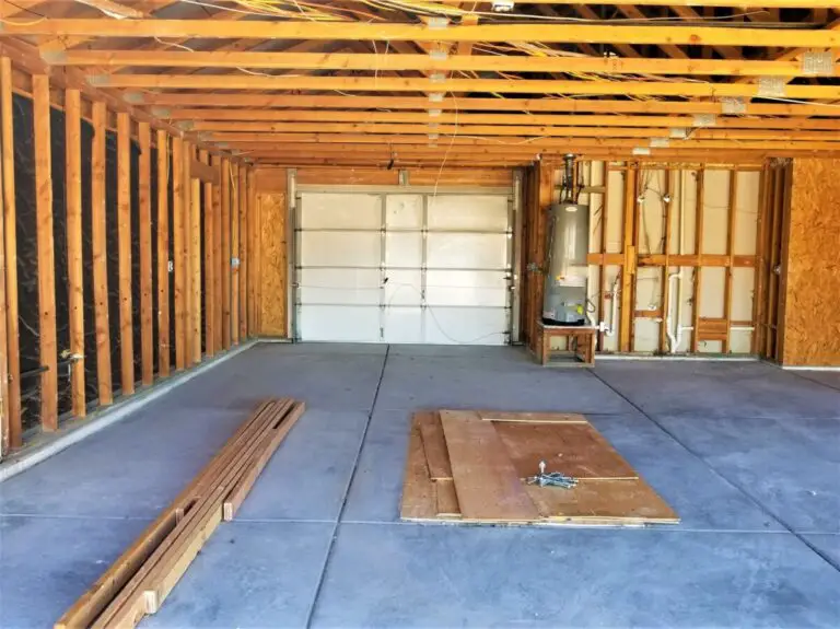 Intérieur de garage non fini avec charpente en bois apparente, poutres de plafond visibles, porte de garage fermée, chauffe-eau et planches de bois sur le sol en béton.