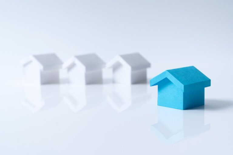 Une maison en papier bleu se dresse au premier plan, tandis que trois maisons en papier blanc sont floues à l'arrière-plan sur une surface blanche brillante.