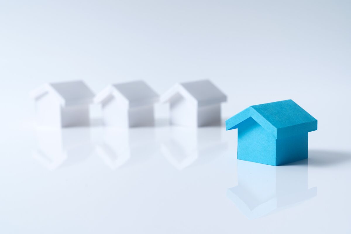Une maison en papier bleu se dresse au premier plan, tandis que trois maisons en papier blanc sont floues à l'arrière-plan sur une surface blanche brillante.