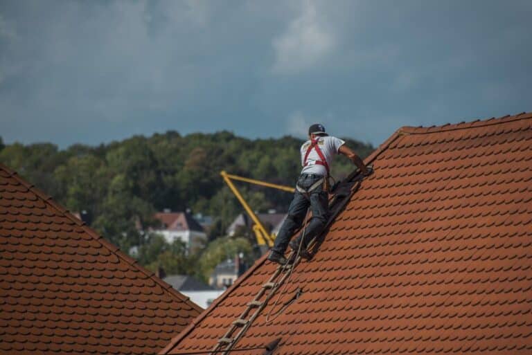 Un travailleur portant un harnais de sécurité se tient debout sur un toit en tuiles, utilisant une échelle comme support, avec des arbres et des bâtiments visibles en arrière-plan.