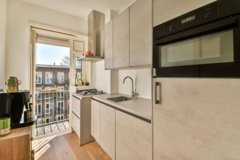 Cuisine moderne et compacte avec des armoires en bois clair des appareils encastrés une cuisinière à gaz et une porte menant à un petit balcon avec vue sur les bâtiments voisins