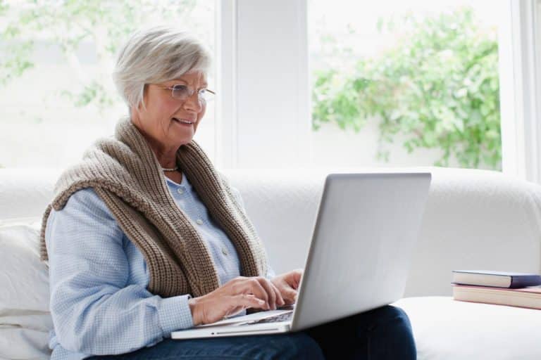 Une femme âgée portant des lunettes et un foulard est assise sur un canapé souriant tout en utilisant un ordinateur portable deux livres sont empilés à côté d elle des fenêtres lumineuses se trouvent à l arrière plan