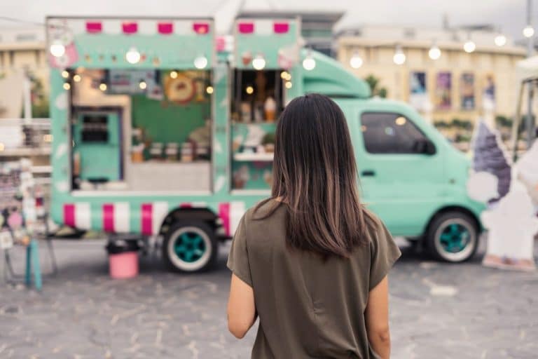 Une femme aux cheveux mi longs se tient face à un food truck turquoise et rose garé en plein air sur une aire pavée