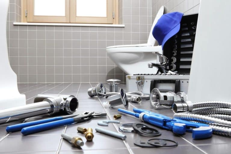 Une salle de bains avec des outils et des pièces de plomberie éparpillés sur le sol, une boîte à outils et un bouchon bleu à côté des toilettes, ce qui laisse supposer des travaux de réparation en cours.