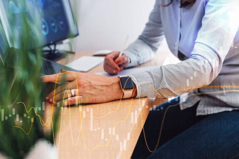 Une personne portant une smartwatch travaille à un bureau utilisant un ordinateur portable et prenant des notes sur papier avec des graphiques de données et des tableaux superposés au premier plan