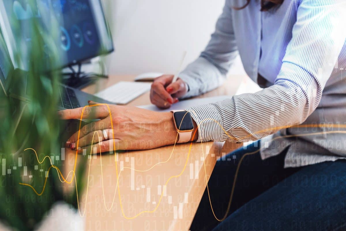 Une personne portant une smartwatch travaille à un bureau utilisant un ordinateur portable et prenant des notes sur papier avec des graphiques de données et des tableaux superposés au premier plan