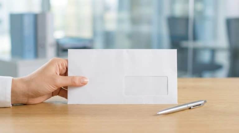 Une main tient une enveloppe blanche avec une fenêtre placée sur un bureau en bois à côté d un stylo argenté dans un environnement de bureau
