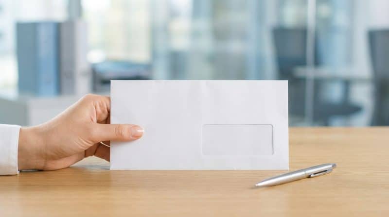 Une main tient une enveloppe blanche avec une fenêtre placée sur un bureau en bois à côté d un stylo argenté dans un environnement de bureau